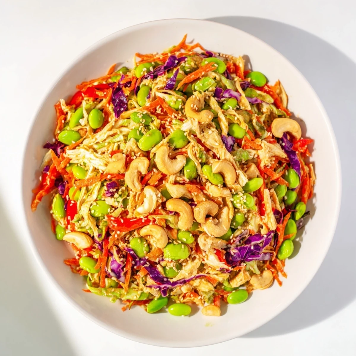 A close-up of Edamame Crunch Chicken Salad in a white bowl, highlighting crisp greens, red bell pepper strips, and golden roasted cashews drizzled with ginger dressing.