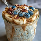 A jar of High-Protein Peanut Butter Overnight Oats topped with sliced bananas, blueberries, and chopped roasted peanuts.