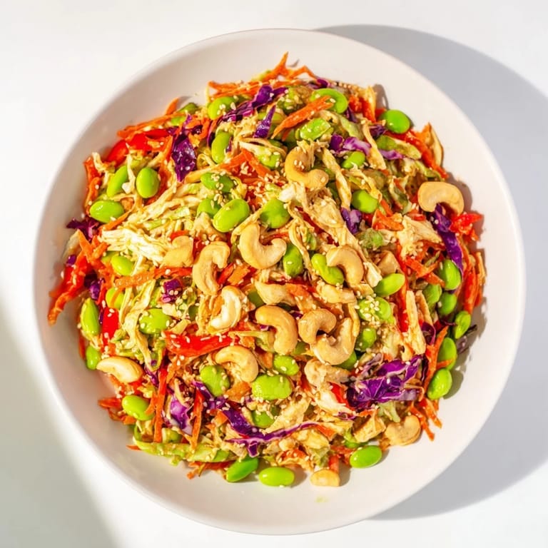 A close-up of Edamame Crunch Chicken Salad in a white bowl, highlighting crisp greens, red bell pepper strips, and golden roasted cashews drizzled with ginger dressing.